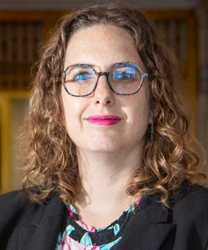 Headshot of Veronica Aveis, a white woman with green eyes and shoulder  length curly brown hair wearing a floral print top, black blazer, and teal  tortoise shell glasses. Aveis smiles directly at the camera.