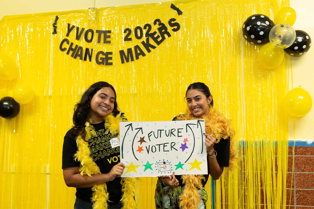 Two teenage girls of Southeast Asian descent with dark brown hair smile holding up a sign pointing to them that says “Future Voter” against a background of bright yellow streamers and a YVote 2023 Change Makers” banner