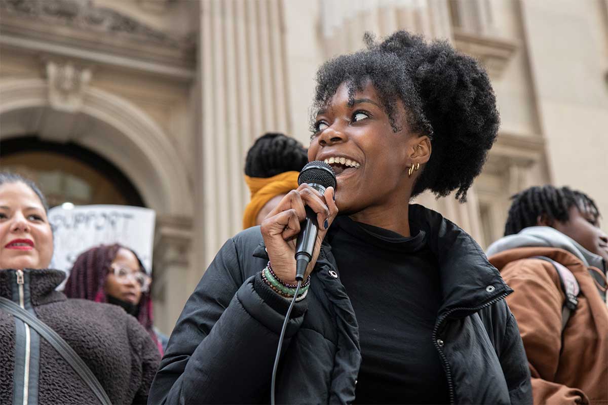 A young woman of color with a black shirt and puffer jacket holds a microphone in her right hand, speaking into it animatedly as she looks out on a crowd of fellow protesters.