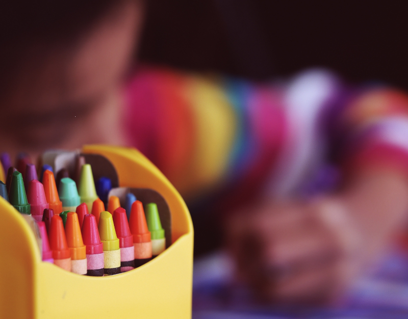 A close-up of a box of colorful crayons. A child, out-of-focus, is seated coloring