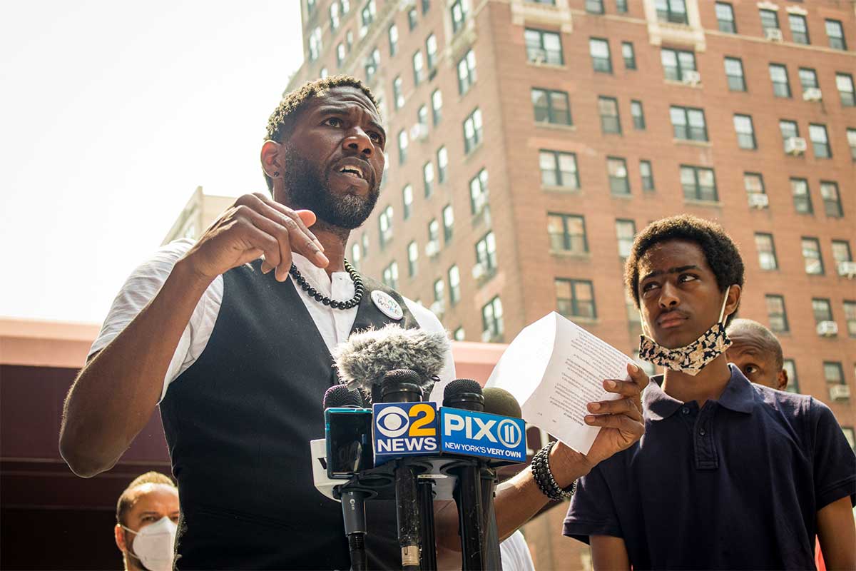 Public Advocate Williams stands in front of a mic stand while speaking to a crowd and  holding a piece of paper in one hand and motioning passionately with the other – a young person standing to his left listens intently as the PA speaks.