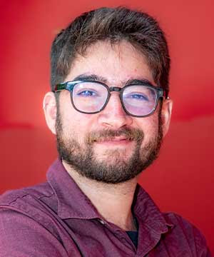 Headshot of David Kahn, a white male with short brown hair, a beard,  black-framed glasses, and a red button-down shirt. David smiles directly at  the camera.