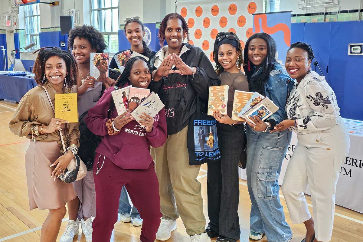 Members of a sorority smile as they hold up books like “The Color Purple” and “The Hill We Climb.” One holds her hands up in a triangle shape to represent Delta Sigma Theta.