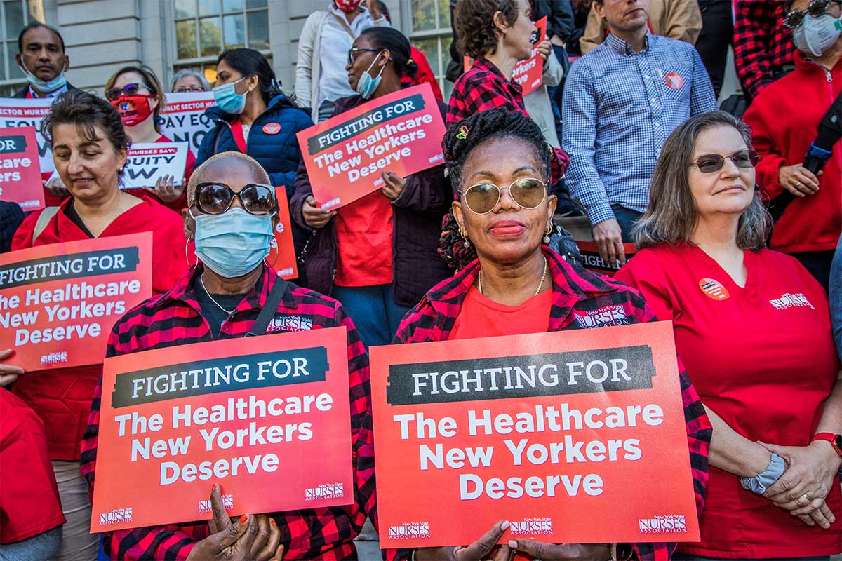 Members of the New York State Nurses Association, many wearing red, gather for a rally on the steps of City Hall. Two women of color look directly at the camera, holding red “Fighting for the Healthcare New Yorkers deserve” signs.