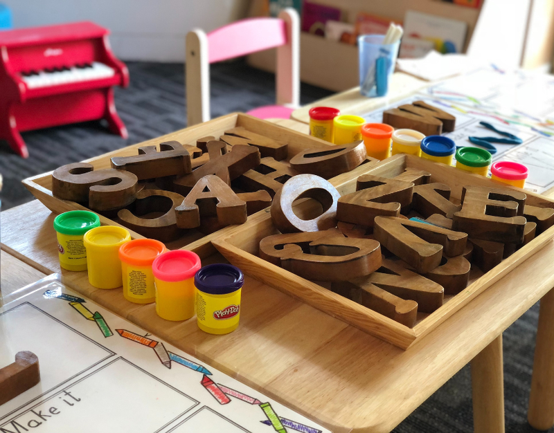 A table in a pre-school with play-doh, large wooden block letters, and coloring pages