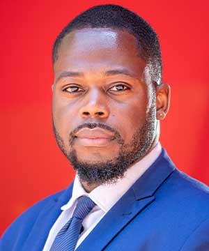 Headshot of Norris Johns. A black man with a low haircut and beard wearing a blue suit and tie stands in front of a red background, looking at the camera.