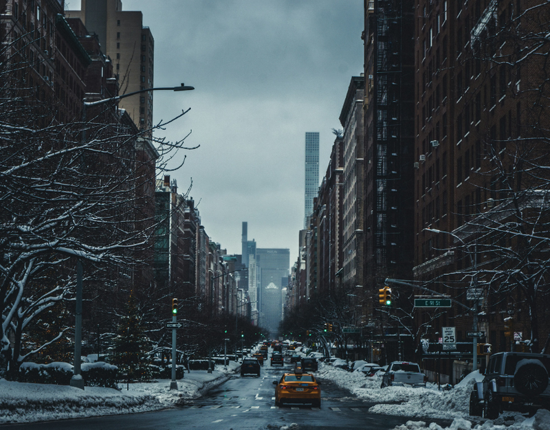 A yellowcab drives down a snowy street with large residential buildings on the Upper West Side