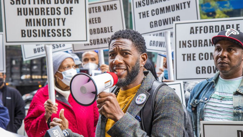 Public Advocate Williams speaks into a megaphone with an outraged expression at a rally. Attendees hold signs with messages like, “Affordable Insurance Keeps the Community Going.”