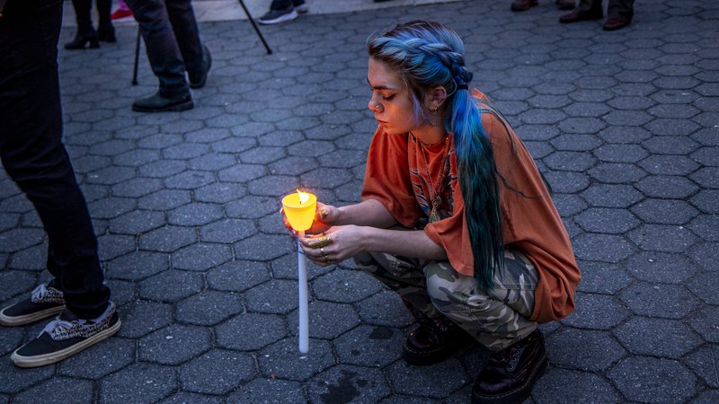 A young woman with braided blue hair and camo pants kneels in Union Square at a gun violence vigil, holding a lit candle