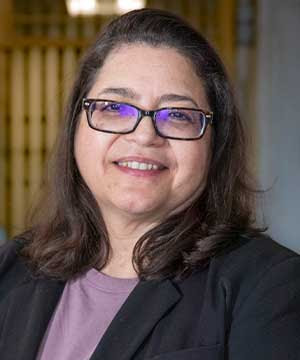 Headshot of Rosie Mendez, a light-skinned Latina with brown hair, eyes, and  glasses. Mendez is smiling directly at the camera with her hands clasped  together in front of her.