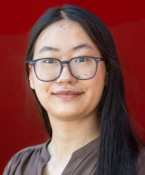 Headshot of Kai Min Yu, an Asian woman with black hair and glasses.  Kai Min smiles at the camera with her hair positioned on one side in front of a red background.