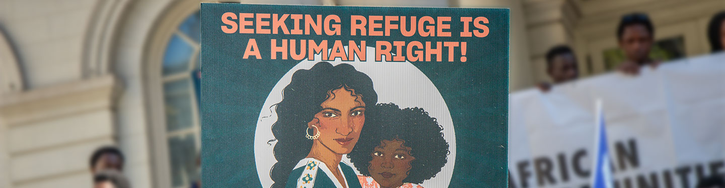 A hand holds a sign stating "Seeking Refuge is a Human Right" amidst a crowd.