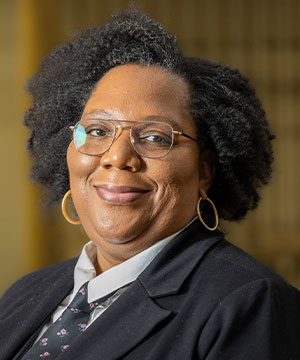 Headshot of Stephanie Gutierrez, a black woman with dark brown curly hair and gold glasses, wearing a black blazer, black vest, tie, and a white blouse smiling directly at the camera.
