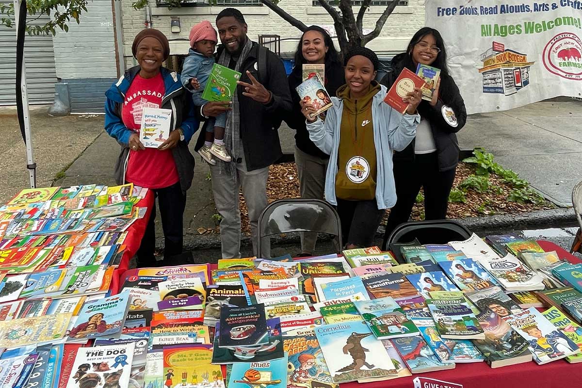 The Public Advocate, holding his daughter as a toddler and children’s book, poses with volunteers at a Brooklyn Book Bodega table. Dozens are books for children of all ages are displayed.