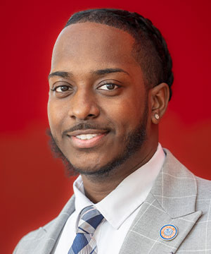 Jeff Senatus, Special Assistant smiling with red background with grey suit and OPA lapel pin
