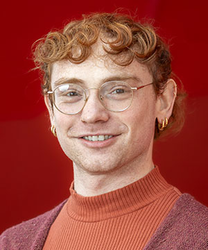Headshot of Ariel Munczek Edelman, a white genderqueer person with short red curly hair and glasses. They smile at the camera in front of a red background.