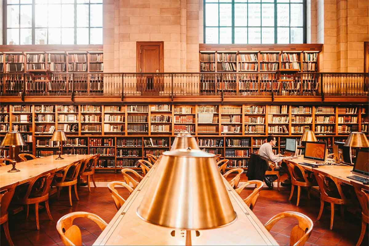 Photo of the interior of the main branch of the New York Public Library with wall full of books and several desks with lamps.