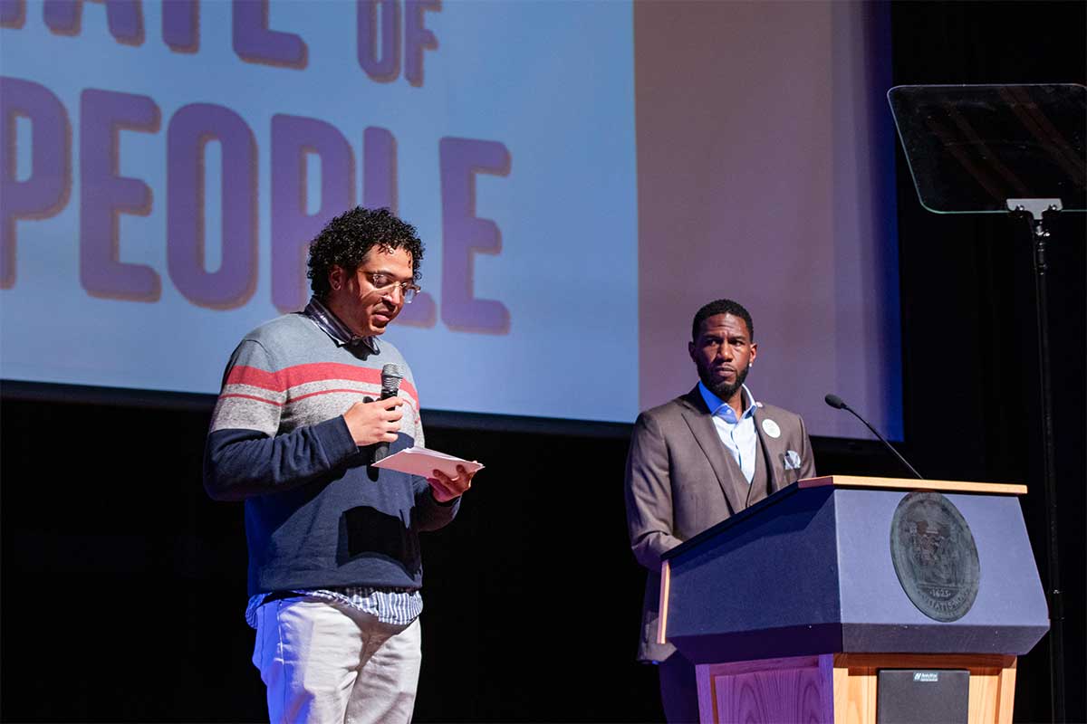 The Public Advocate, with a serious expression, listens to Felix Guzman speak from a notecard on stage at the 2024 State of the People.