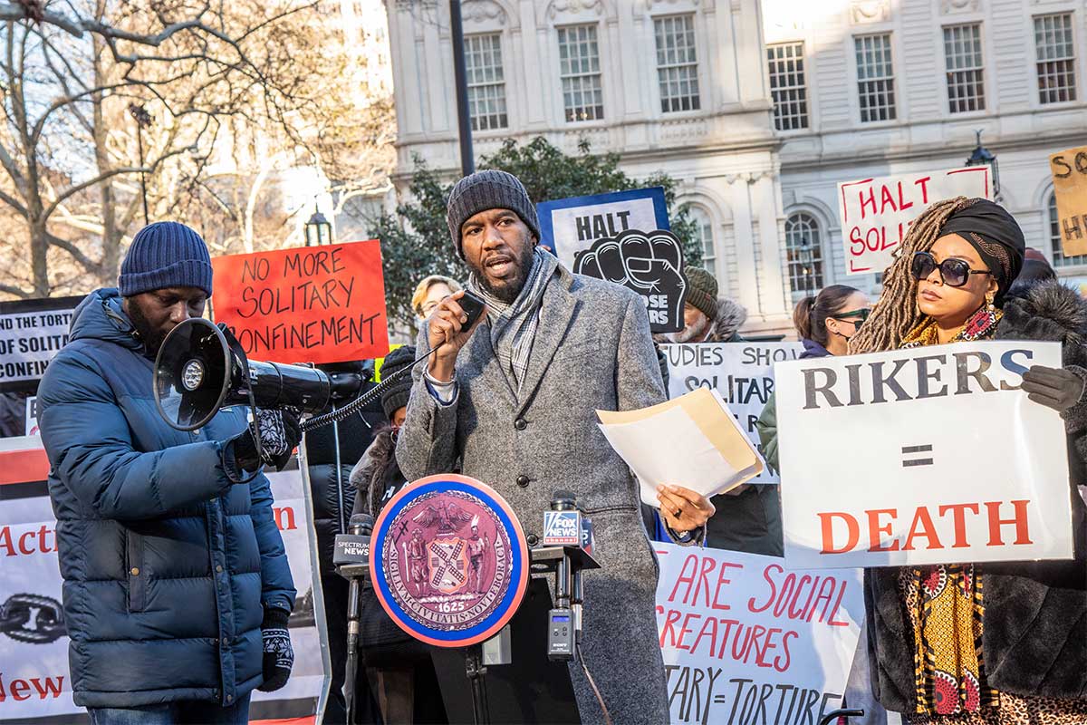 The Public Advocate speaks into a megaphone in City Hall Park, with advocates holding signs with messages like, "No more solitary confinement"