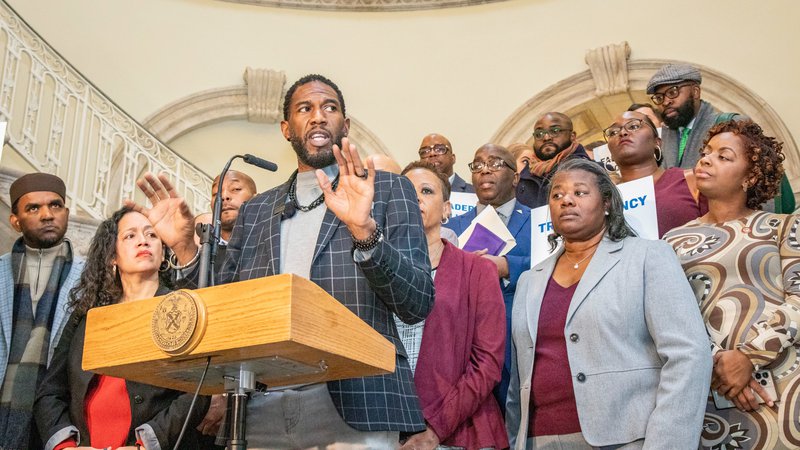 The Public Advocate stands behind a wooden lectern in the City Hall rotunda. Behind him are Speaker Adrienne Adams, Council Member Alexa Aviles, and a group of faith leaders