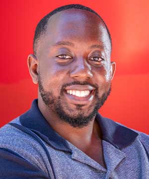 Black male, with a medium stocky build smiling as he folds his arms wearing a navy blue cuban shirt.