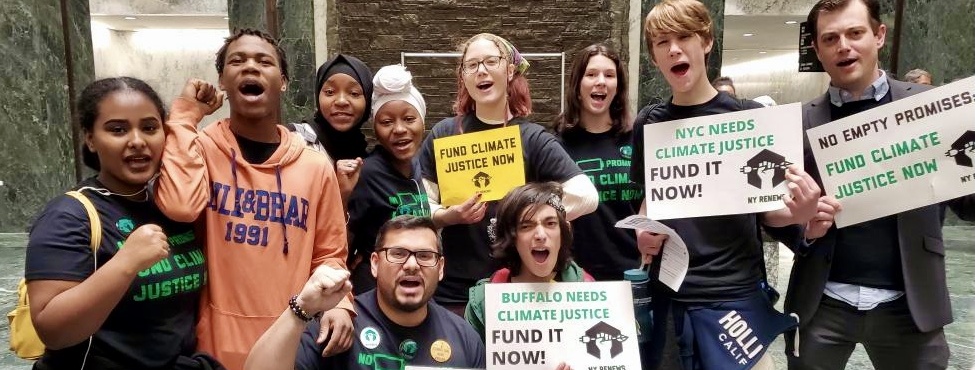Deputy Public Advocate Kashif Hussain and Steve Fox pose with youth activists in Albany capitol building. Many raise fists and hold signs that read, 'NYC Needs Climate Justice. Fund it now!'