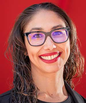 Headshot of Lupe Hernandez, an indigenous Venezuelan woman with long wavy brown hair and black glasses. Lupe is wearing a black dress and blazer while smiling directly at the camera. 