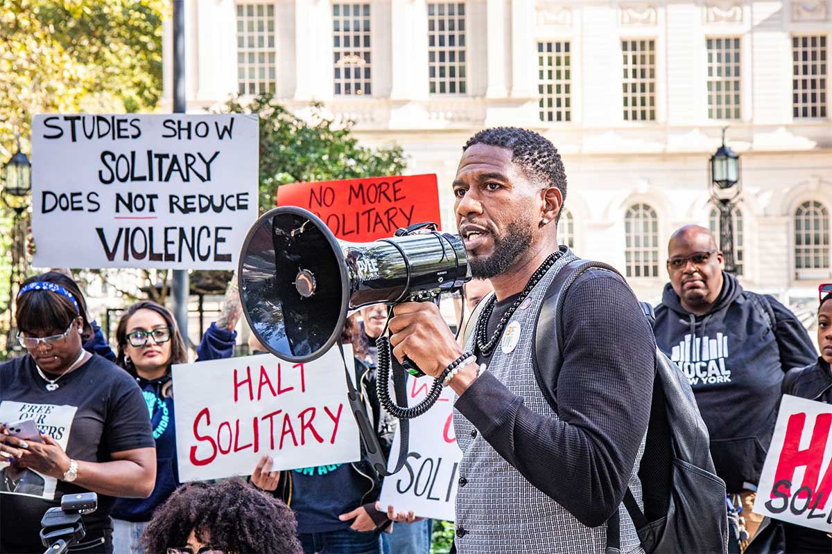 Public Advocate Jumaane D. Williams holding a megaphone at a halt solitary confinement rally. Several people holdings signs in support stand behind him.