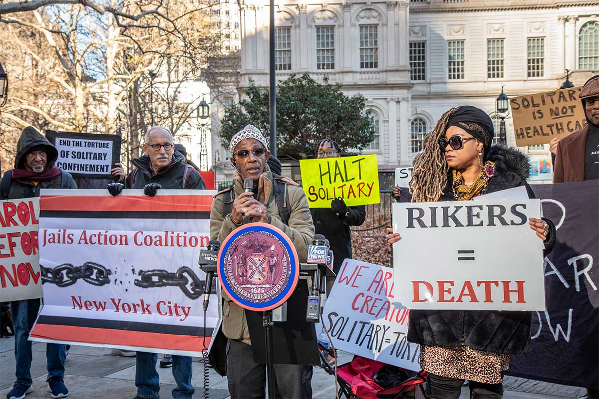 Victor Pate speaks at a rally in City Hall Park with a serious expression. Advocates hold signs with messages like, “Rikers = Death” and “Solitary is not healthcare.”