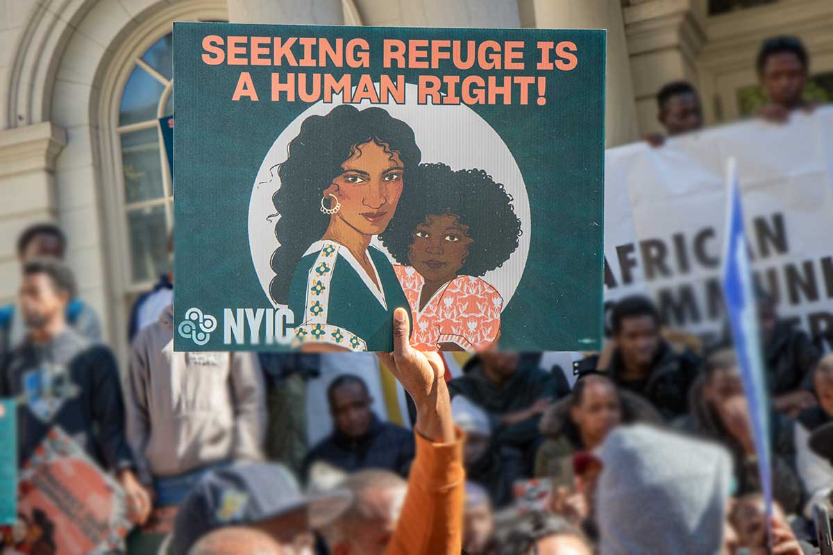 A hand holds a sign stating "Seeking Refuge is a Human Right" amidst a crowd. 