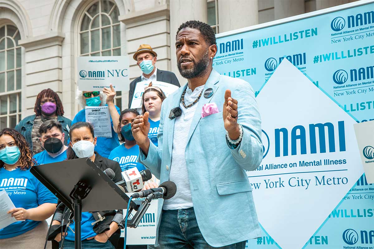 The Public Advocate speaks at a rally in front of City Hall in front of a step-and-repeat for The National Alliance on Mental Illness New York City Metro. Advocate and elected officials listen behind, holding #MentalHealthMatters signs.