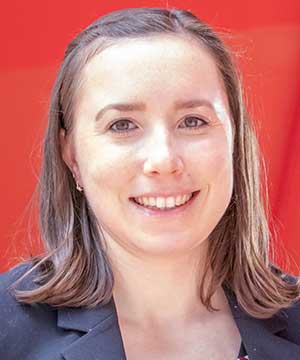 Headshot of Gwen Saffran, a young white woman with shoulder-length brown hair wearing a navy blue top and black blazer. She stands against a red background with her hands on her hips and is smiling at the camera.