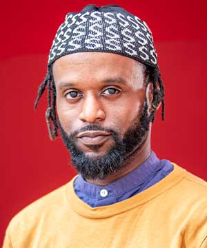 Headshot of Raymond Sheares, a Black man with locks and beard. Ray stares glaringly into the camera.