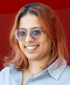Headshot of Nishat Chowdhury, a brown woman with shoulder-length dyed red hair and heart-shaped glasses. Nishat's head is slightly tilted, and she smiles directly at the camera.