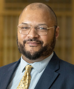 Johnnie Davis is a Black man with short black hair. He is wearing a dark blue suit, a light blue shirt, and gold patterned tie with gold-rimmed glasses.